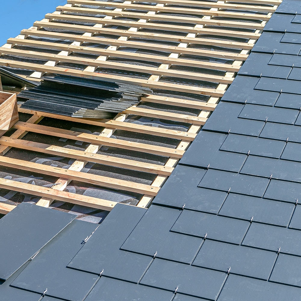 Pose de tuiles d'ardoise sur des liteaux en bois lors d'une rénovation de toiture sous un ciel bleu.